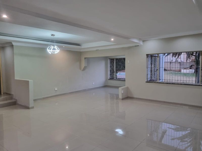 A spacious, open-plan living area featuring polished porcelain floor tiles, a recessed ceiling with modern lighting, and large windows with security bars.