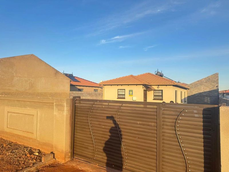 Front view of the property featuring a secure metal sliding gate, perimeter wall, and a tiled roof house with solar water heating.