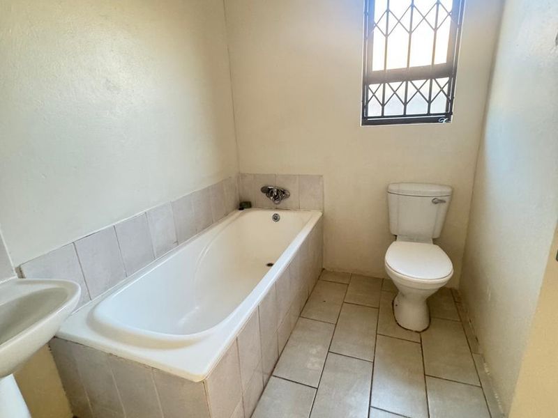 A functional bathroom featuring a bathtub, pedestal basin, and toilet, finished with tiled flooring and a window with a security gate.