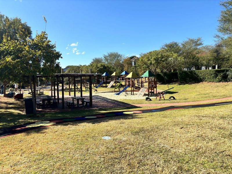 A well-maintained communal playground area featuring various play structures, a shaded pergola with seating, and landscaped garden surroundings.