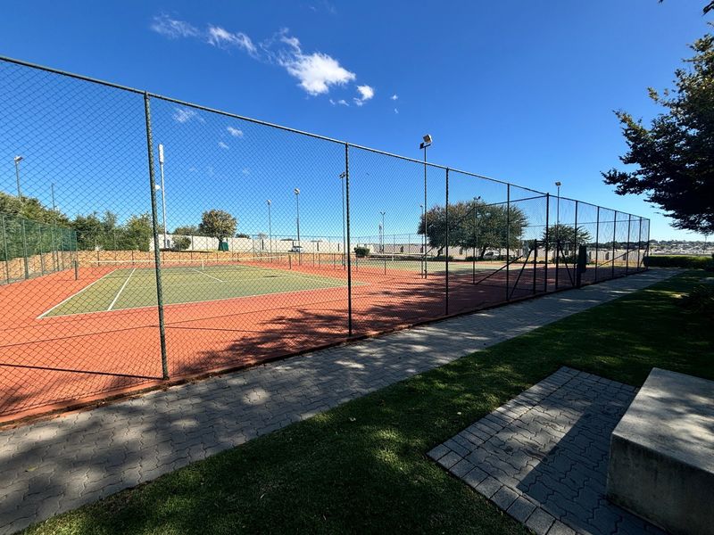 A well-maintained outdoor tennis court facility featuring perimeter chain link fencing, floodlighting, and adjacent paved walkways within the apartment complex.