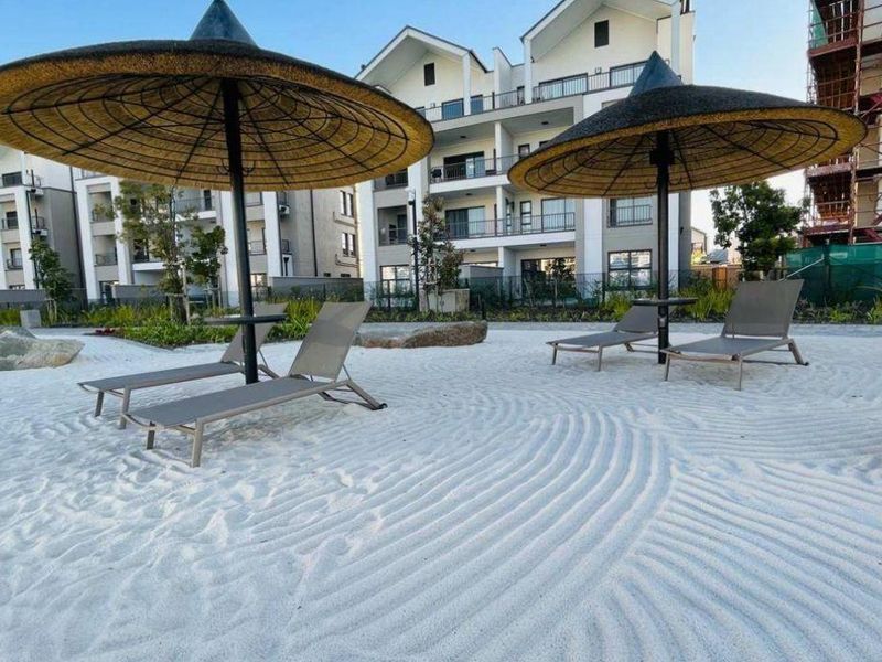 A well-maintained communal outdoor leisure area featuring a sand beach aesthetic with landscaped surroundings and modern apartment buildings in the background.