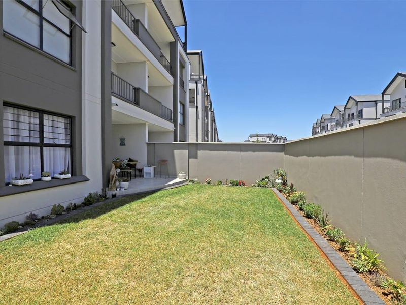 A private rear garden featuring a well-maintained lawn, landscaped garden beds, and a paved patio area adjacent to the apartment building.