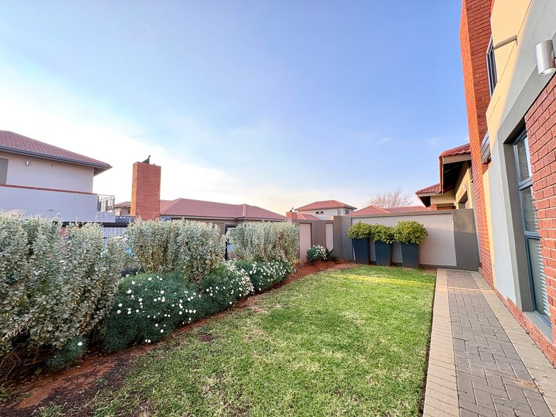 A well-maintained rear garden area featuring a manicured lawn, brick paved pathway, and established perimeter landscaping against a boundary wall.
