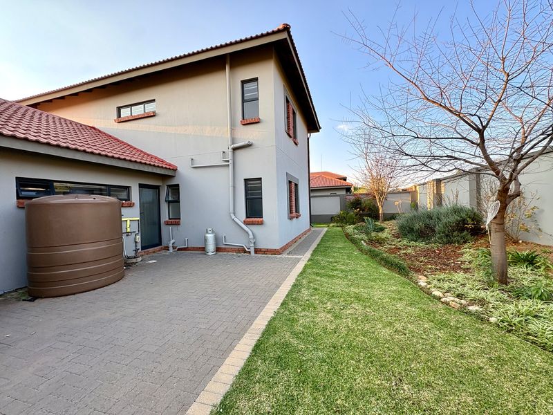 Rear exterior view of the property featuring a paved patio area, water storage tank, and a well-maintained garden with lawn and established trees.