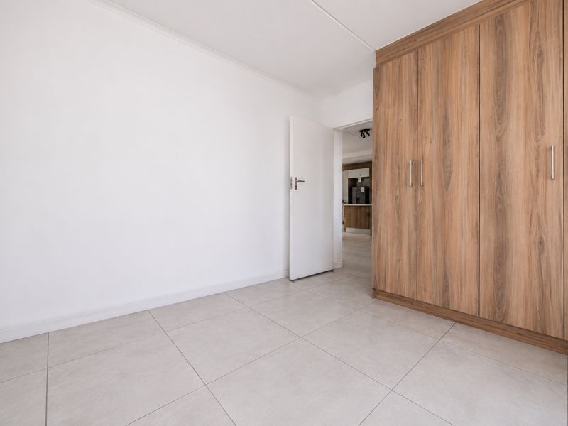 A bright, empty bedroom featuring modern light-toned floor tiles and a large, built-in wooden wardrobe with ample storage space.