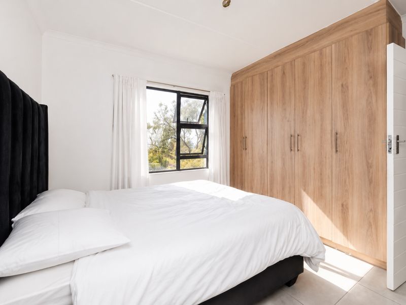 A bright bedroom featuring modern wood-finish built-in cupboards, tiled flooring, and an aluminium window frame overlooking greenery.