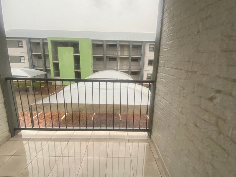 A covered balcony featuring tiled flooring and a metal balustrade, overlooking the apartment complex parking area.