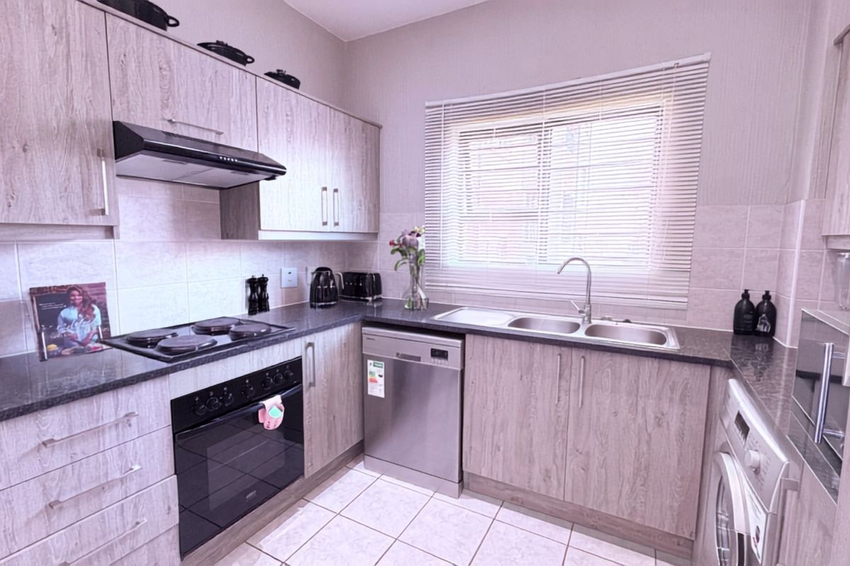 A well-maintained kitchen featuring light wood-grain cabinetry, dark laminate countertops, integrated electric hob and oven, and a double bowl sink with tiled flooring.