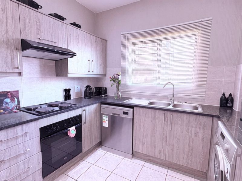 A well-maintained kitchen featuring light wood-grain cabinetry, dark laminate countertops, integrated electric hob and oven, and a double bowl sink with tiled flooring.