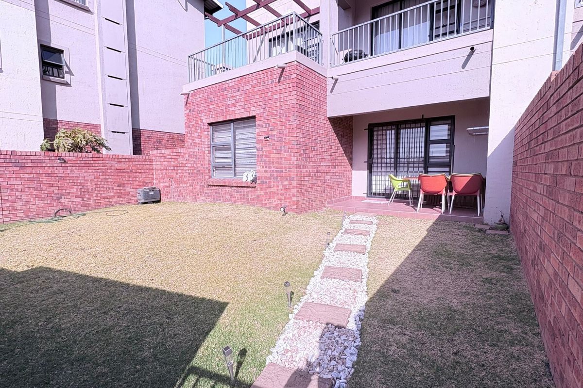 A private rear garden featuring a lawn, stone pathway, and a paved patio area with access to the apartment via sliding glass doors.