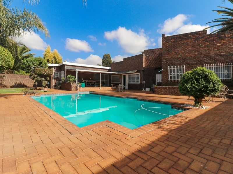 A spacious outdoor pool area featuring a swimming pool, extensive brick paving, and a covered patio attached to the face brick residence.