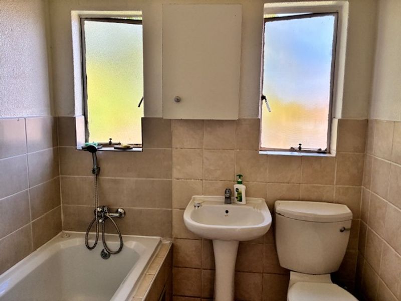 A functional bathroom featuring a bathtub, pedestal basin, toilet, and wall-mounted cabinet, finished with neutral wall tiling and dual frosted windows.