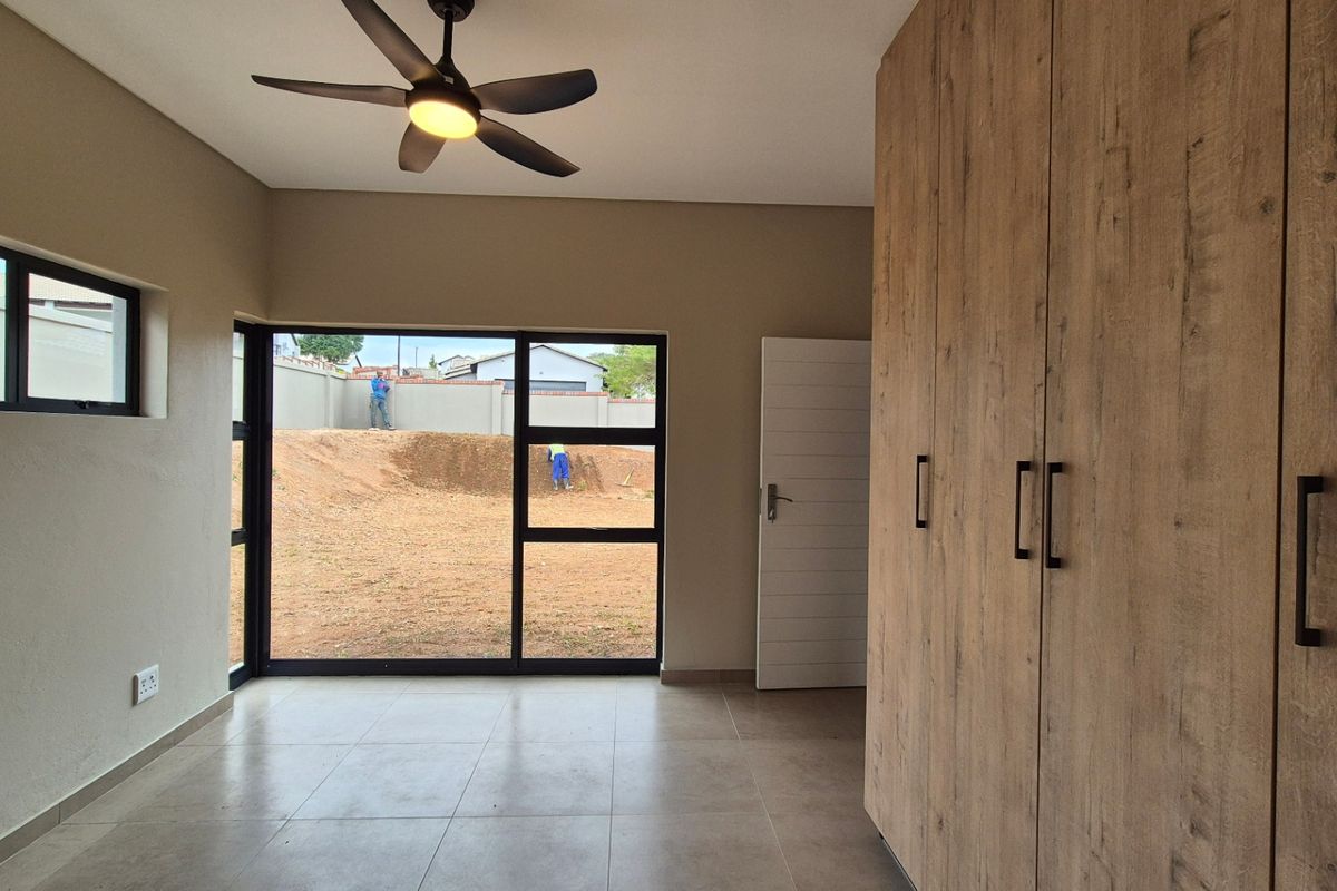 A bright, modern bedroom featuring neutral tiled flooring, built-in wood-finish cupboards, a ceiling fan, and large windows overlooking the rear yard.