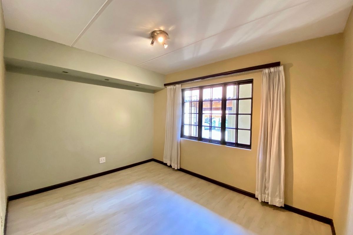 A bright, empty bedroom featuring light-toned laminate flooring, a recessed ceiling bulkhead, and a large grid-pattern window with neutral wall finishes.