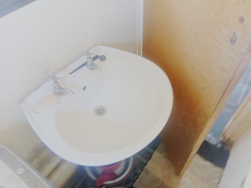 A dated bathroom pedestal basin featuring dual-handle taps, showing signs of wear on the surrounding wall surfaces.