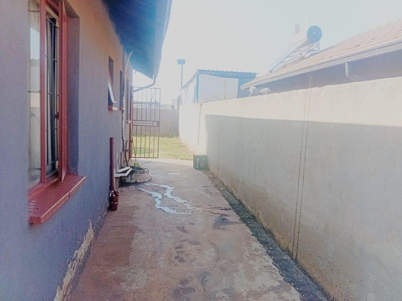 A narrow rear exterior side passage featuring a concrete walkway, boundary wall, and metal security gate, showing signs of wear and weathering.