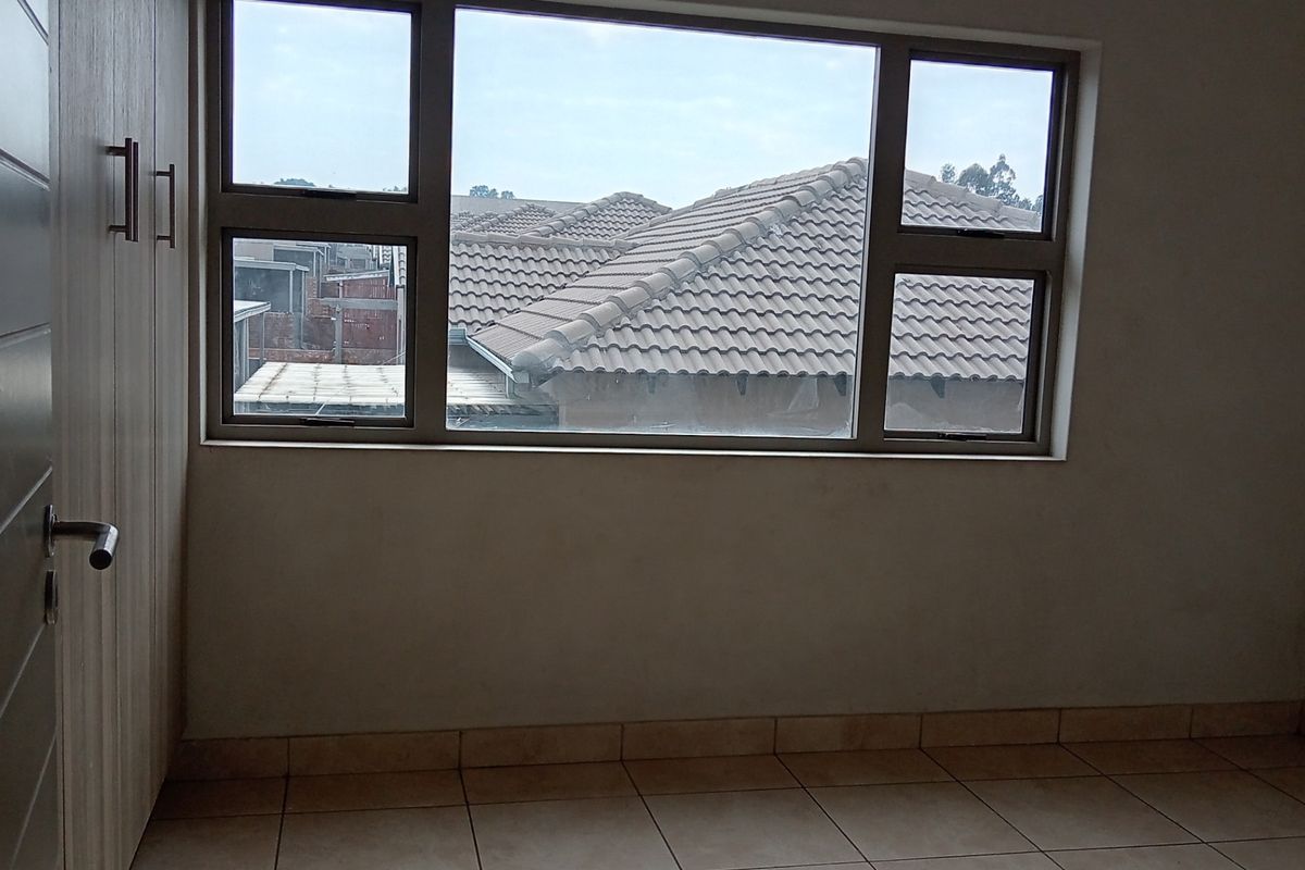An empty bedroom featuring neutral tiled flooring, built-in cupboards, and aluminium-framed windows with a view of the surrounding townhouse complex.