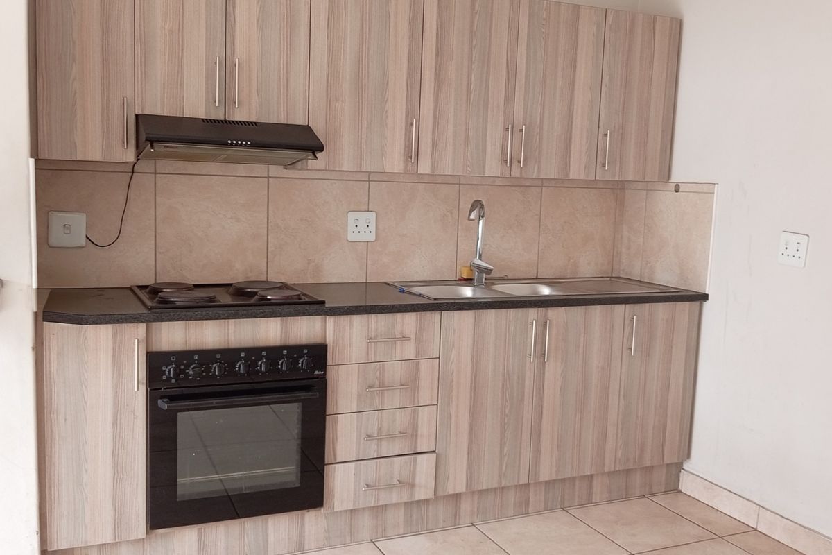 A clean, functional kitchen featuring light wood-grain cabinetry, built-in oven and hob, tiled backsplash, and neutral floor tiles.