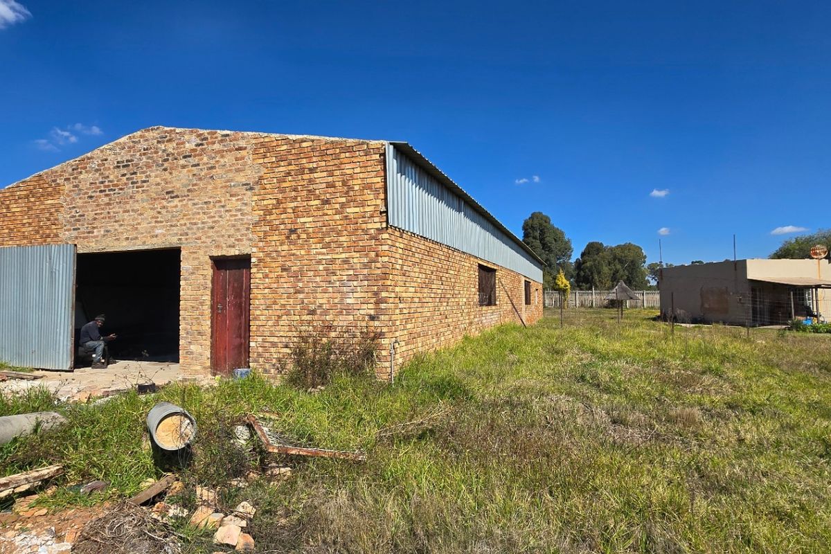 Exterior view of a large brick barn structure with corrugated metal roofing, situated on a rural property with perimeter fencing.