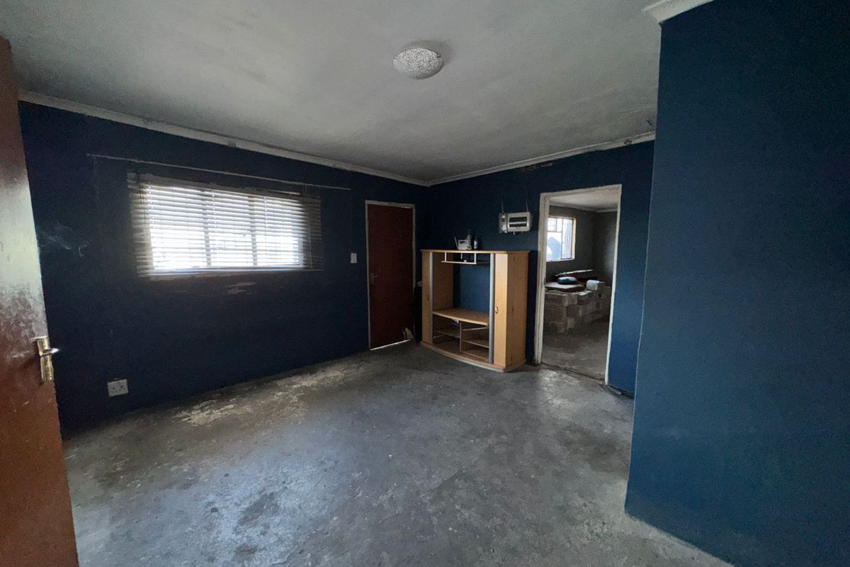 An empty living room featuring dark blue walls, exposed concrete flooring, and a window with venetian blinds, requiring cosmetic updates and flooring installation.