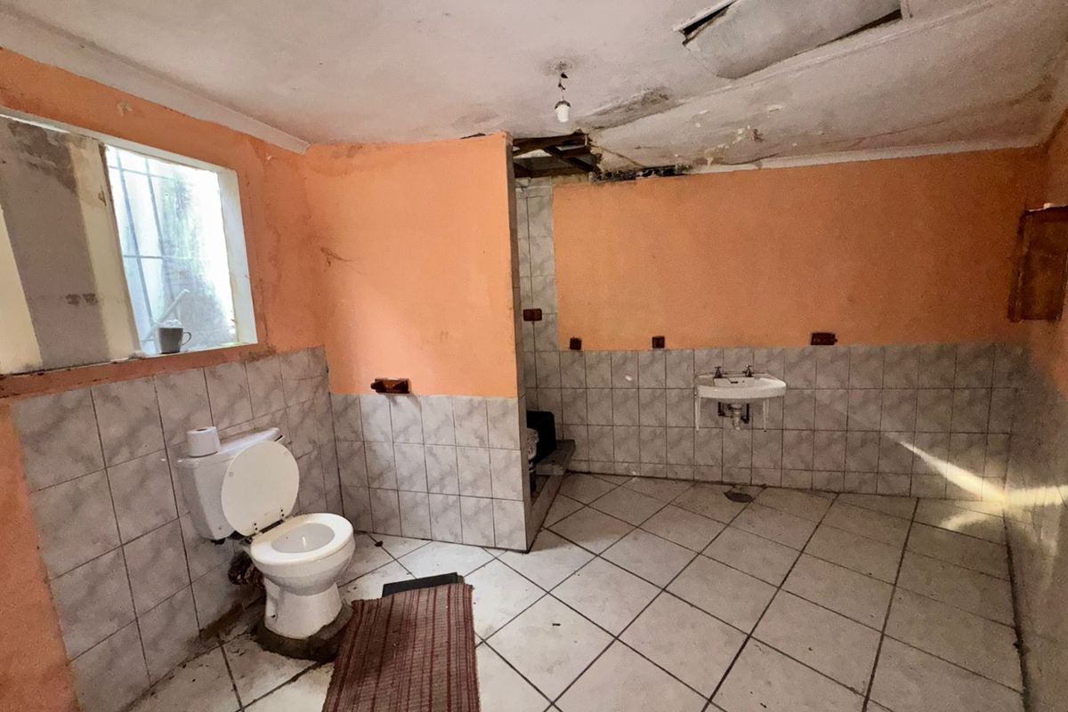 A spacious bathroom requiring significant renovation, featuring a toilet, wall-mounted basin, and tiled finishes with visible ceiling and wall damage.