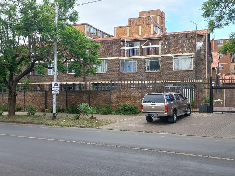 Front exterior view of a multi-story brick townhouse complex featuring a paved driveway, secure perimeter fencing, and gated access.