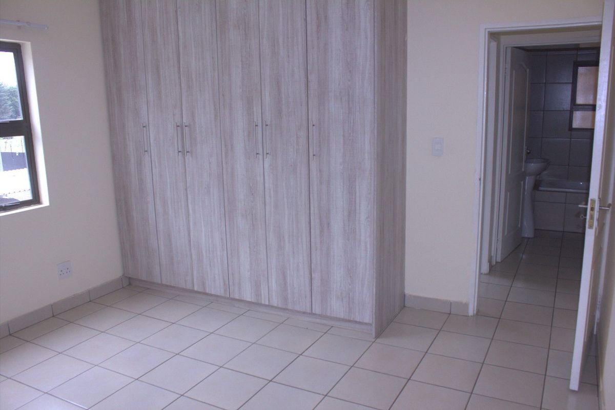 A clean, empty bedroom featuring light wood-grain built-in cupboards, neutral tiled flooring, and a view into the adjacent bathroom.