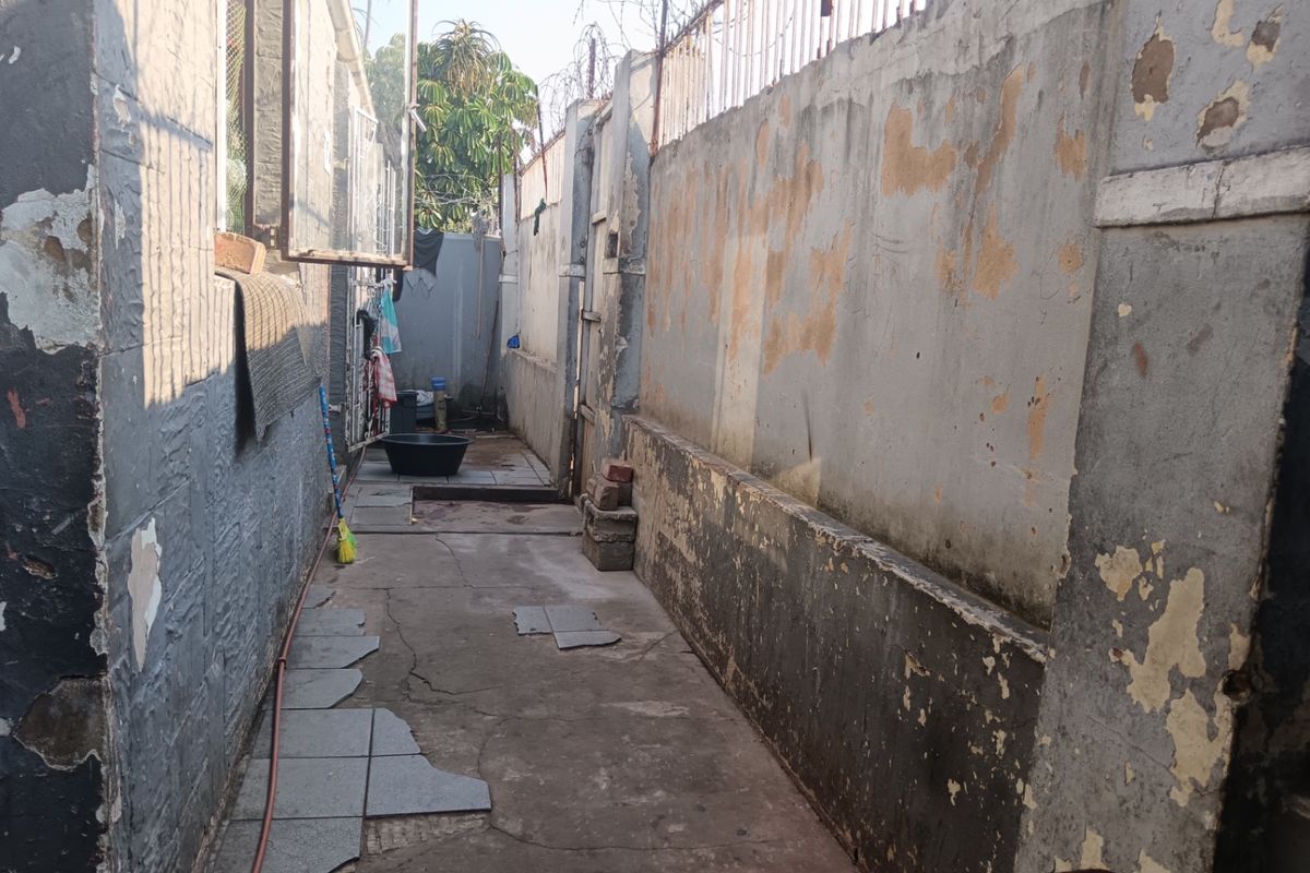 A narrow rear exterior walkway featuring a concrete perimeter wall with security spikes and damaged paved flooring requiring maintenance and repair.
