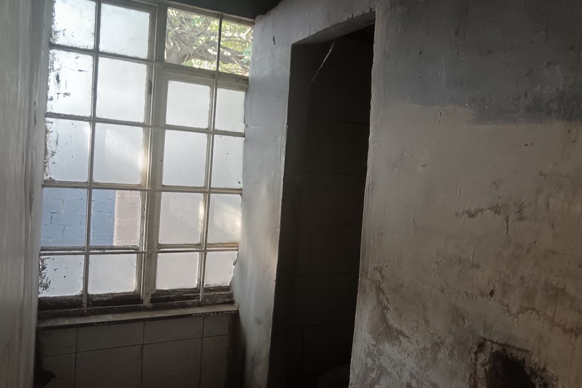 A dilapidated bathroom space requiring significant renovation, featuring a steel-framed window and damaged wall finishes.