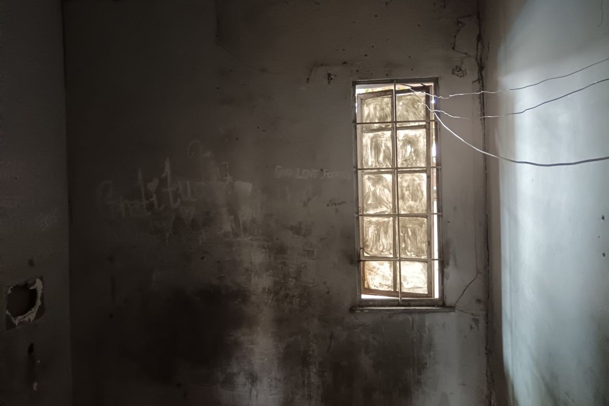 An empty room in poor condition featuring a window with security bars and glass blocks, requiring significant renovation and wall repairs.
