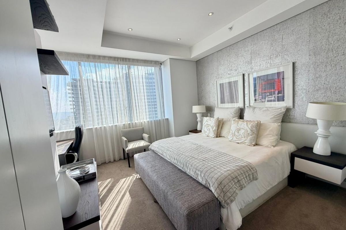 A well-appointed bedroom featuring a textured accent wall, recessed ceiling with downlights, and large windows providing ample natural light.