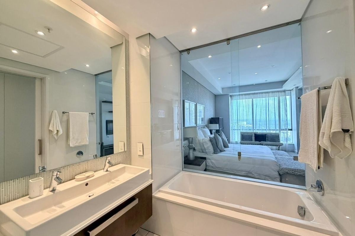 Modern bathroom featuring a double vanity, built-in bathtub, and a glass partition wall overlooking the adjacent bedroom.