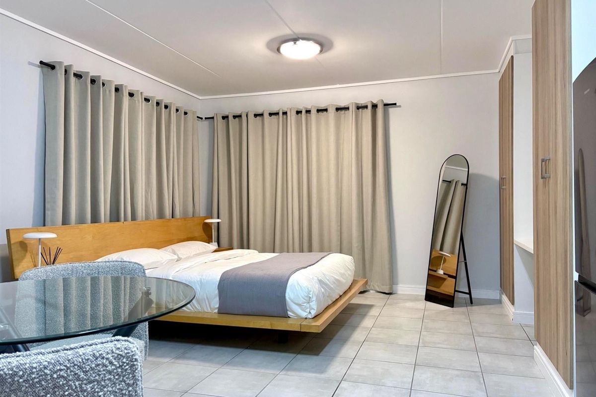 A well-maintained bedroom featuring neutral tiled flooring, built-in wooden cabinetry, and ample natural light through floor-to-ceiling curtains.
