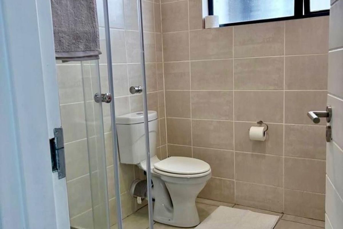 A functional bathroom featuring a glass-enclosed shower, toilet, and neutral wall and floor tiling with an obscured glass window for privacy.