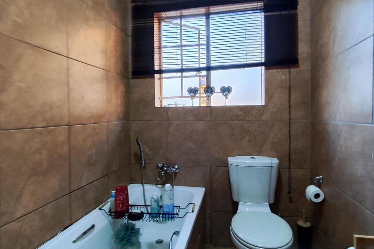 A functional bathroom featuring full-height wall tiling, a built-in bathtub, and a standard toilet, illuminated by a window with fitted blinds.
