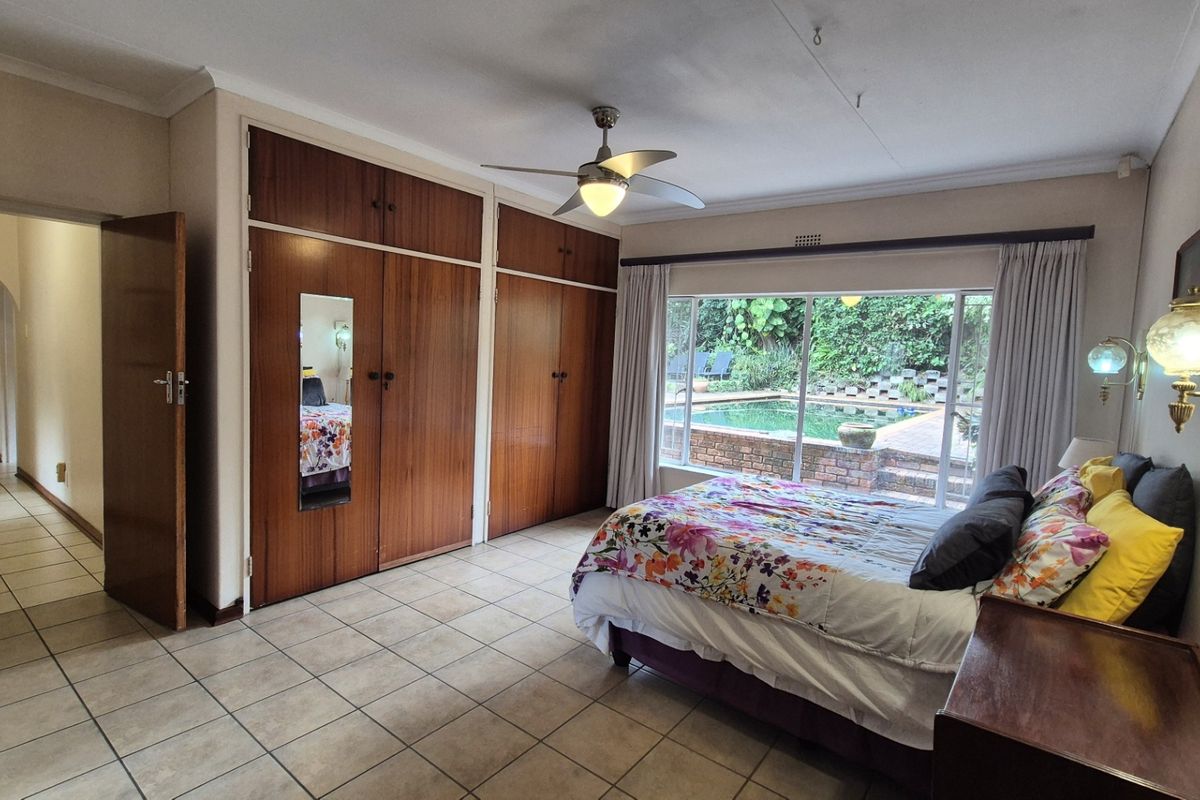 Spacious bedroom featuring built-in wooden cupboards, tiled flooring, a ceiling fan, and large windows offering a view of the outdoor pool area.