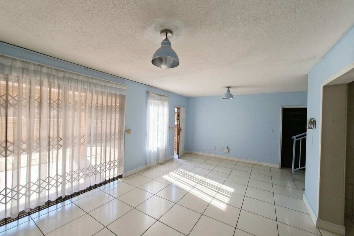 A bright, empty living room featuring tiled flooring, pendant lighting, and a sliding glass door with a security gate leading to the exterior.