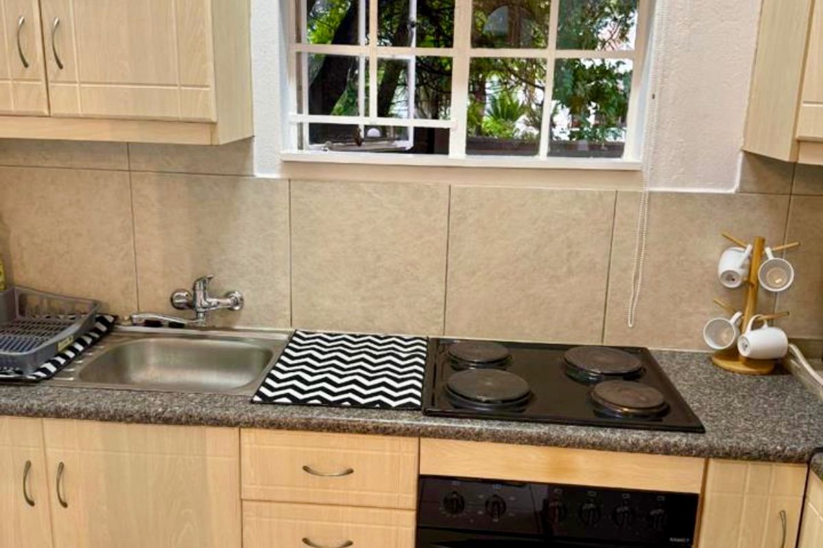 A compact kitchen featuring light wood-finish cabinetry, a built-in oven and hob, tiled backsplash, and a window with security bars and a roller blind.