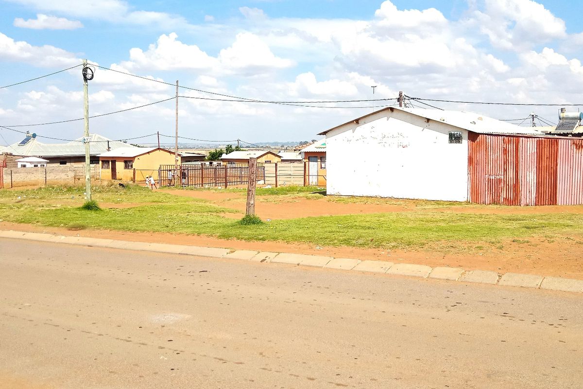 A street-level view of a residential property featuring a corrugated metal structure, solar water heating system, and perimeter fencing.