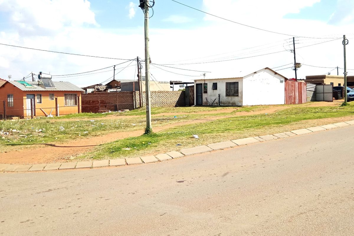 A street-level view of a residential corner plot featuring multiple detached structures, boundary walls, and utility infrastructure in a suburban setting.