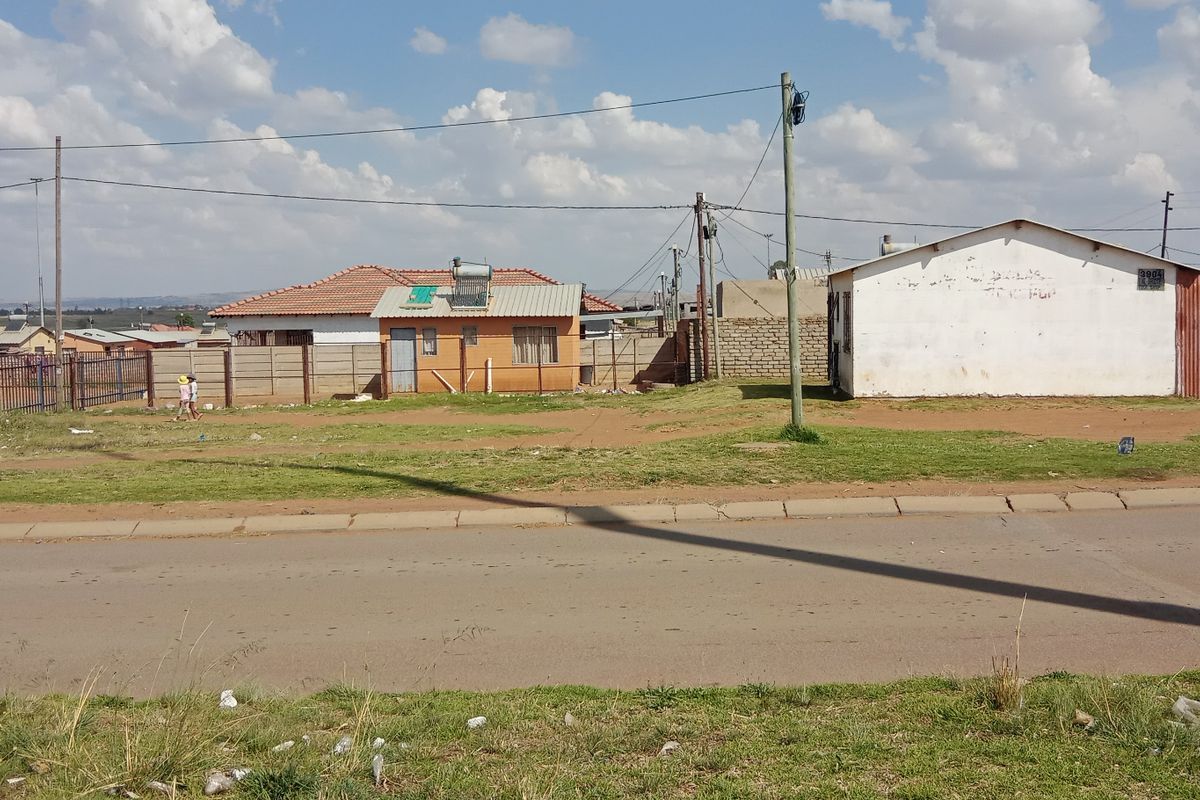 A street-level view of a residential area showing detached houses, perimeter walls, and overhead utility infrastructure under a partly cloudy sky.