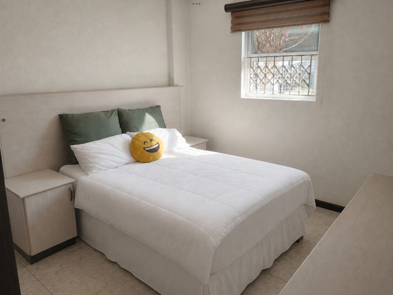 A functional bedroom featuring built-in bedside tables, a matching headboard, tiled flooring, and a window fitted with a security gate and roller blind.