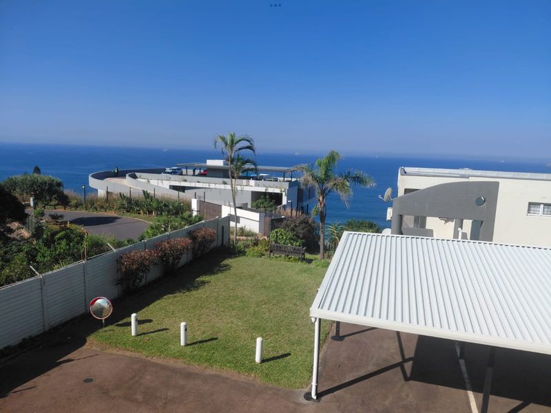 Elevated view overlooking a landscaped garden, paved driveway, carport, and boundary fencing with a scenic ocean backdrop.