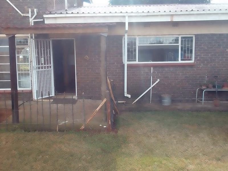 Rear view of a brick house featuring a covered patio area, security gates, and window bars, set against a modest garden space.