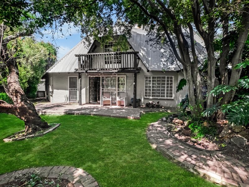 Rear view of the house featuring a corrugated metal roof, a wooden balcony, and a brick-paved patio overlooking a lush garden.