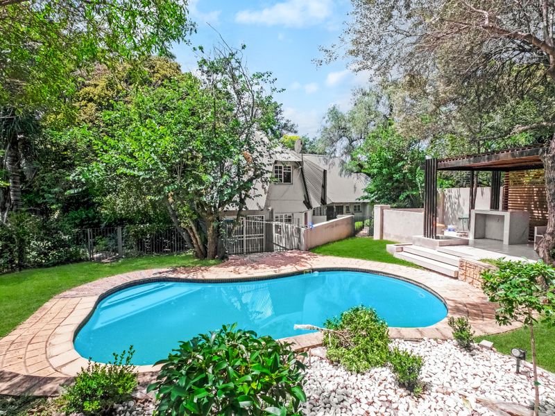 A landscaped backyard featuring a kidney-shaped swimming pool, a covered patio area with a built-in braai, and mature trees.