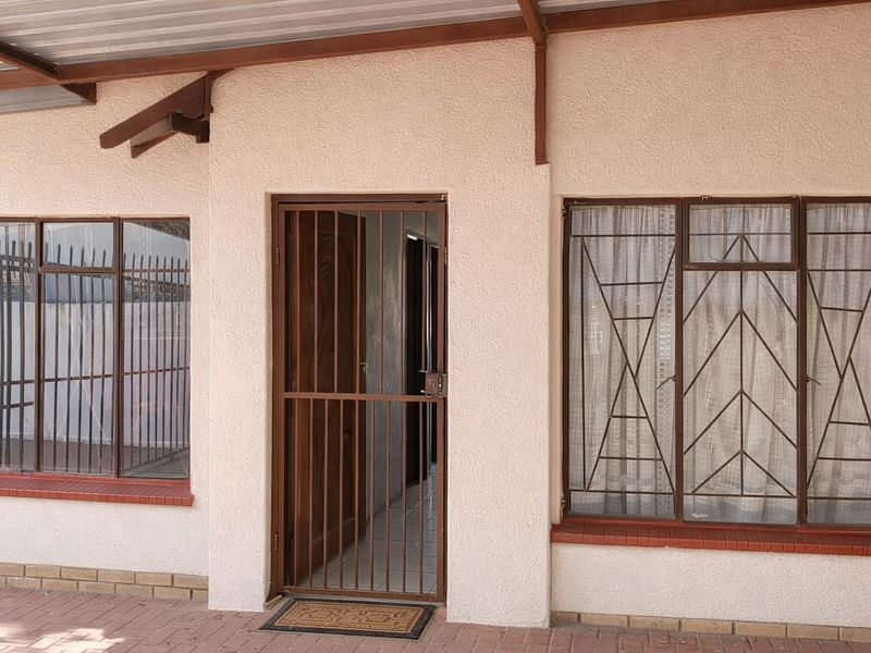 A covered patio area featuring a secure metal entrance gate, brick window sills, and metal-framed windows with security bars.