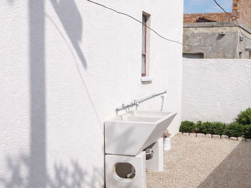 A clean rear exterior area featuring a functional utility sink mounted against a white rendered wall, set on a gravel surface with stone edging.