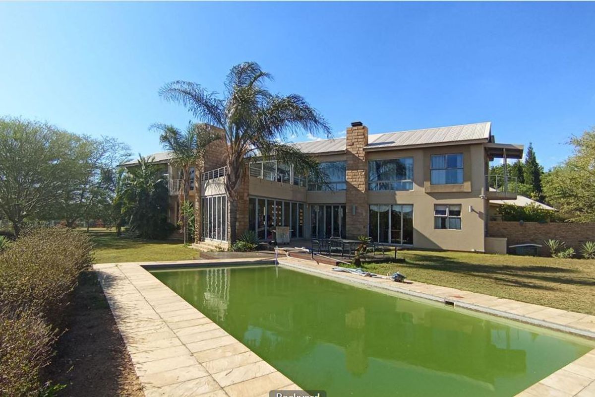 Rear view of a large multi-story house featuring stone cladding, a metal roof, and a swimming pool set within a spacious garden.